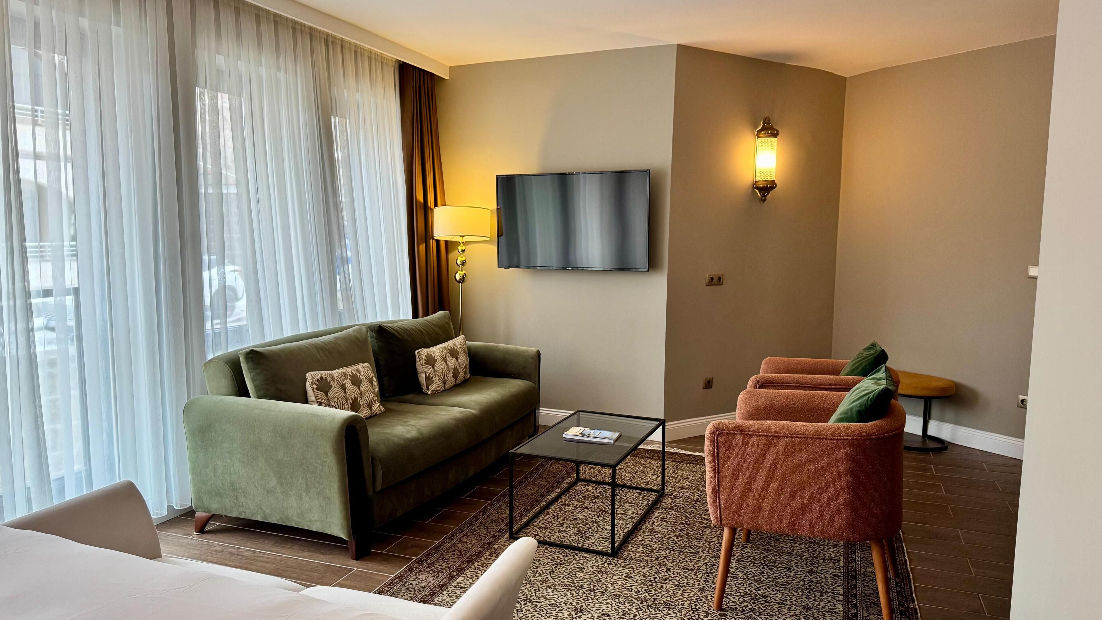 Quarto de hotel com sofá verde, mesa de centro pequena, poltrona vermelha, TV fixada na parede e uma janela grande com cortinas translúcidas.