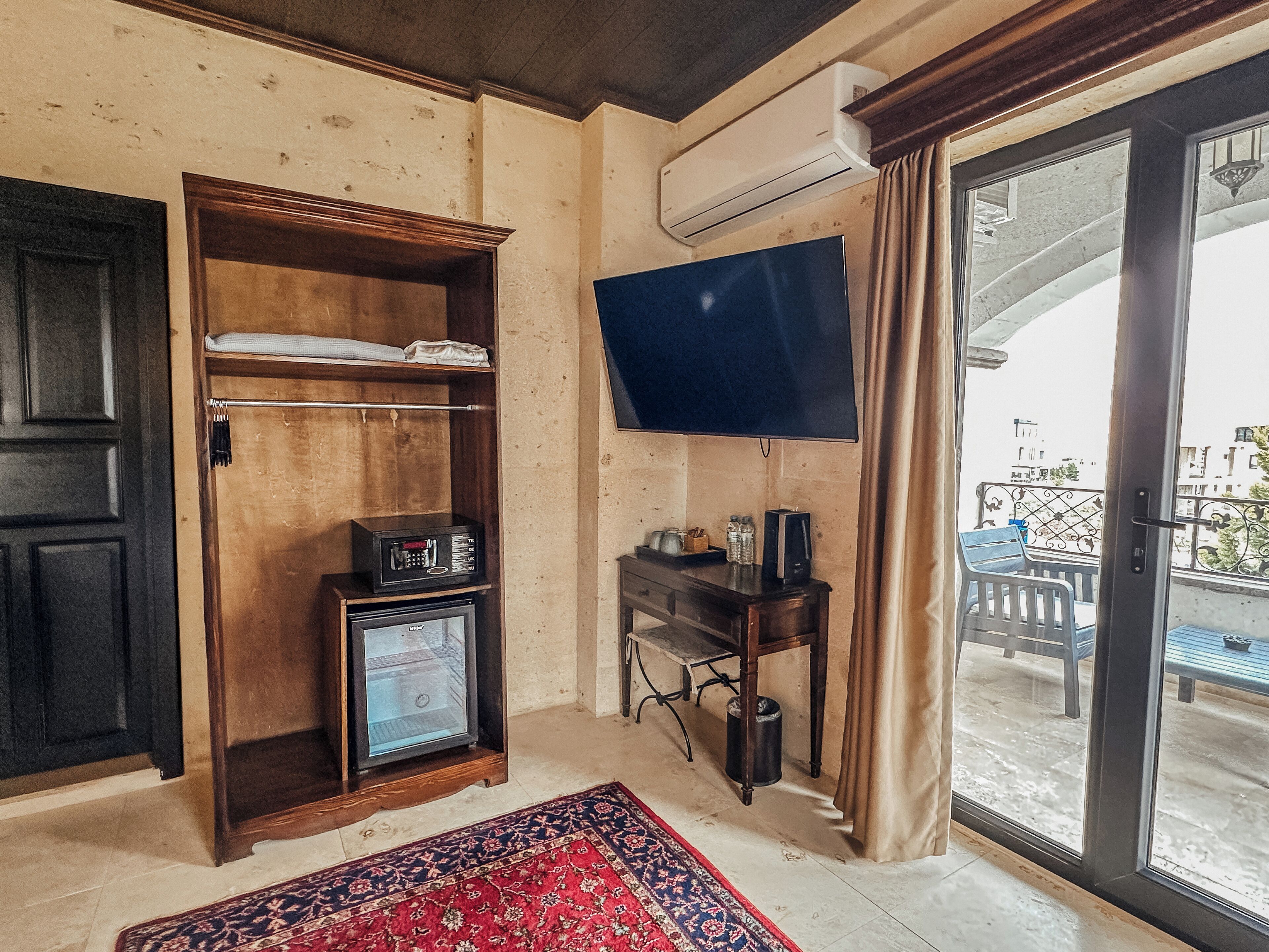 A room with a wooden wardrobe, a television mounted on the wall, a small table with a kettle, and a view of the outside balcony.
