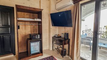 A room with a wooden wardrobe, a television mounted on the wall, a small table with a kettle, and a view of the outside balcony.