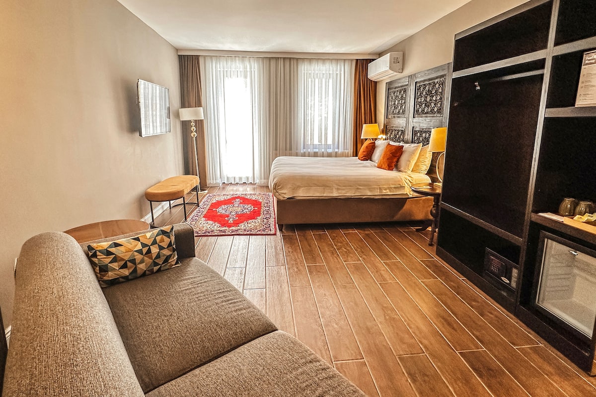A hotel room with a bed, a sofa, a red rug, a floor lamp, and a TV.