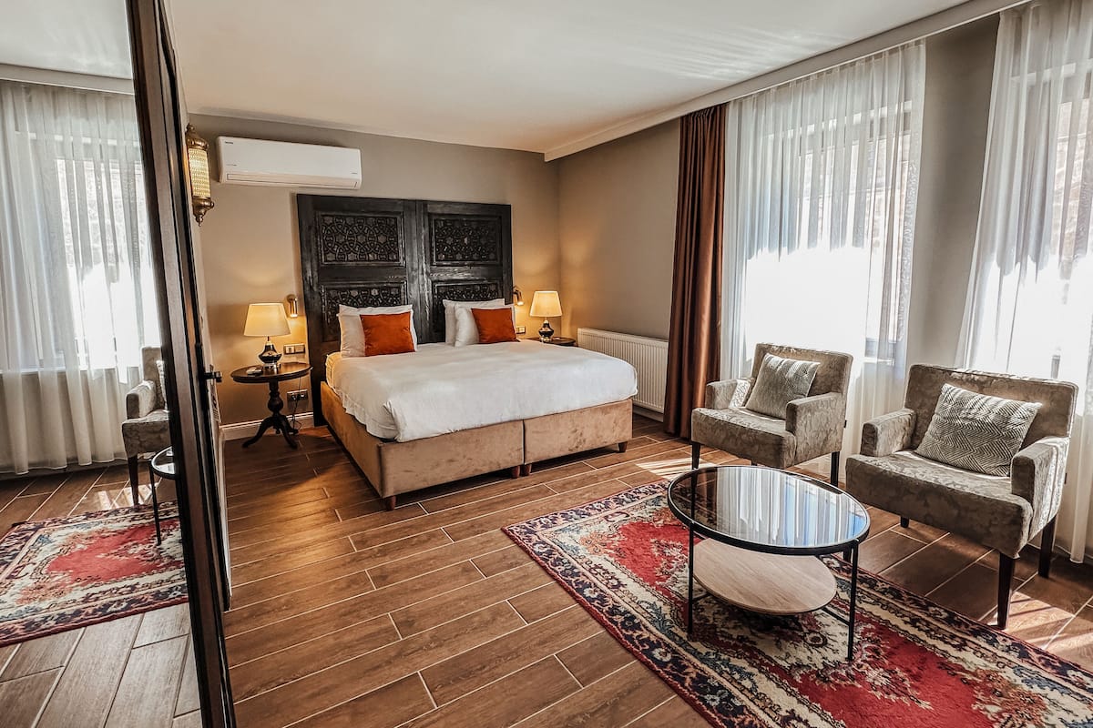 A hotel room with a large bed, two bedside tables with lamps, a round coffee table, two armchairs, and a patterned rug.