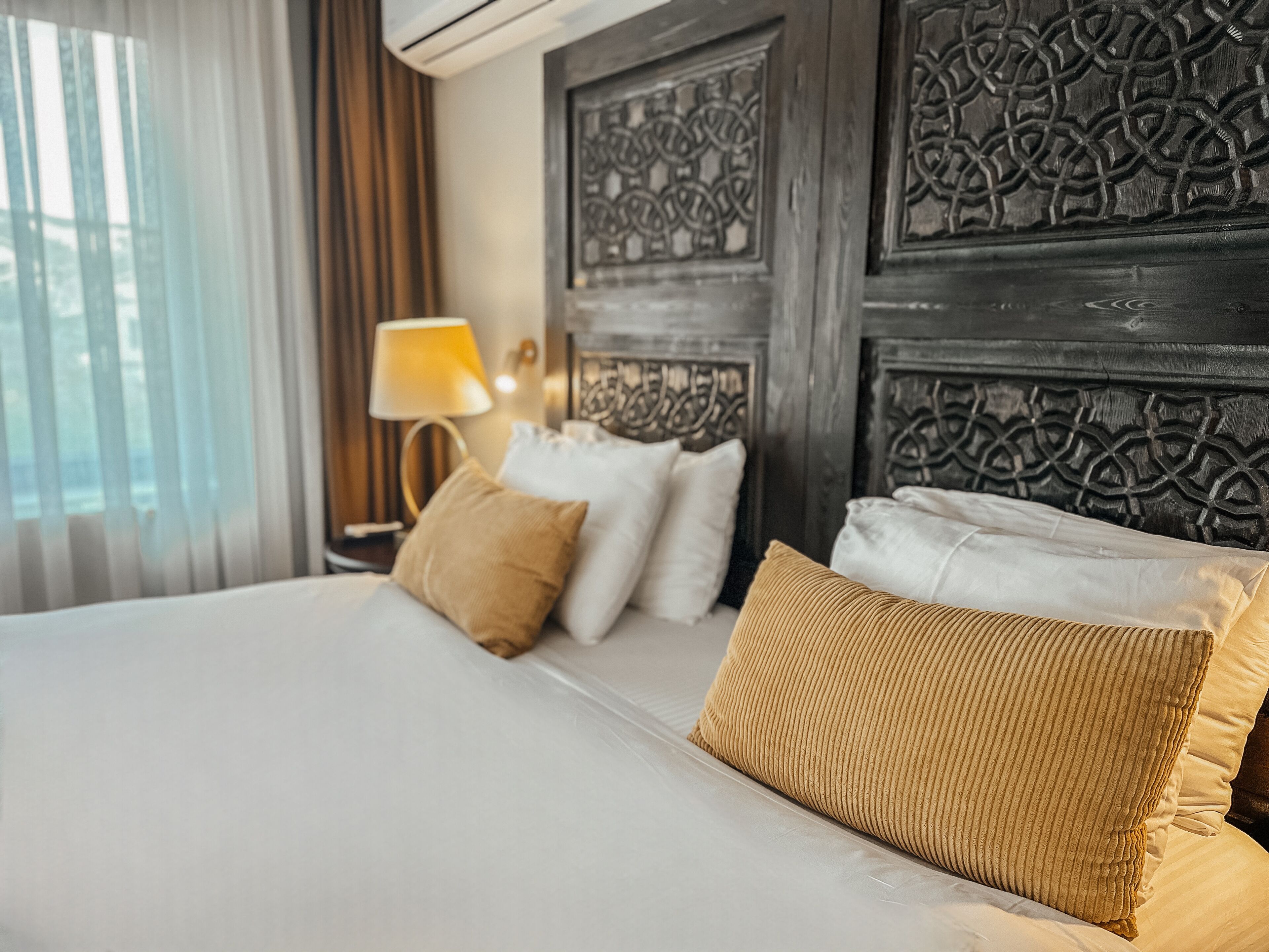 Habitación de hotel con una cama grande, cabecera con elaborados adornos, lámpara de noche y vista por la ventana.