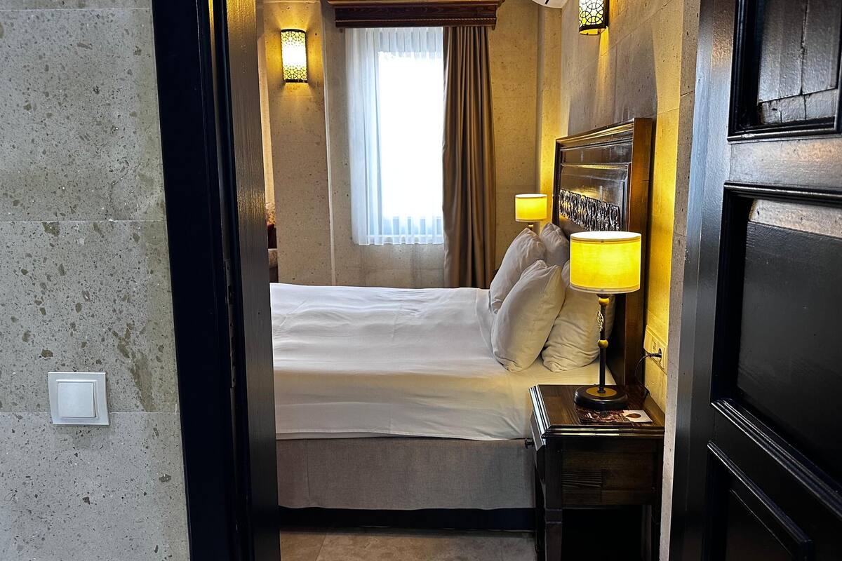 A hotel room with a bed, two bedside lamps, a nightstand, a window with curtains, and a wall-mounted light fixture.