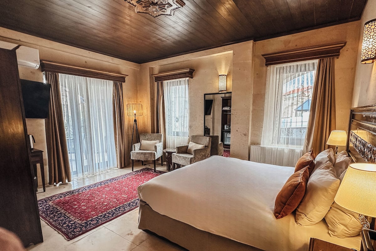 A spacious hotel room with a large bed, two armchairs, a small table, a floor lamp, and a view of the city through large windows.
