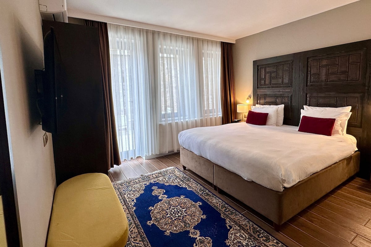 A hotel room with a large bed, a flat-screen TV mounted on the wall, a wooden floor, and a large window with sheer curtains.