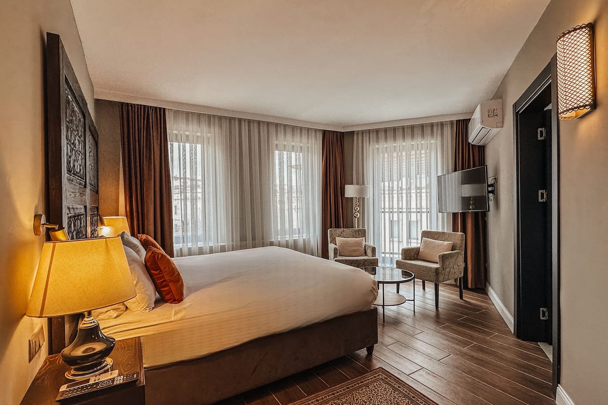 A hotel room with a large bed, a desk with a lamp, two armchairs, a TV, and a window with curtains.