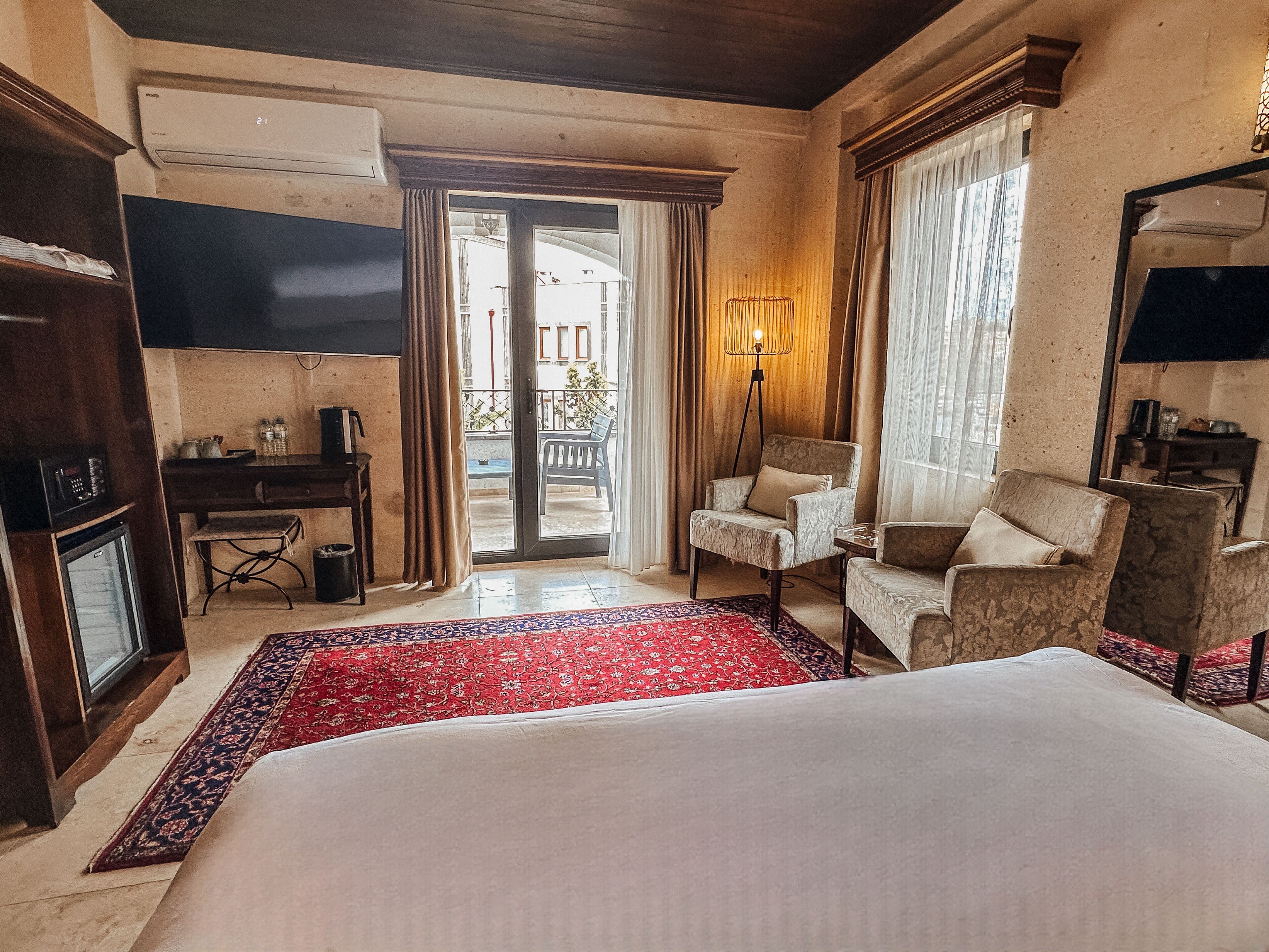 Una camera d'albergo con un letto grande, una scrivania, una sedia, una televisione e un balcone con tende.