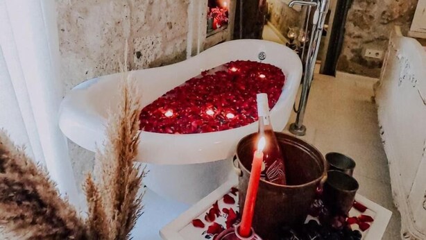 A bathroom with a stone wall, a white bathtub filled with red rose petals, a lit candle, and a plate with grapes and rose petals. - Hoşça kal Alaçatı (Cesme)