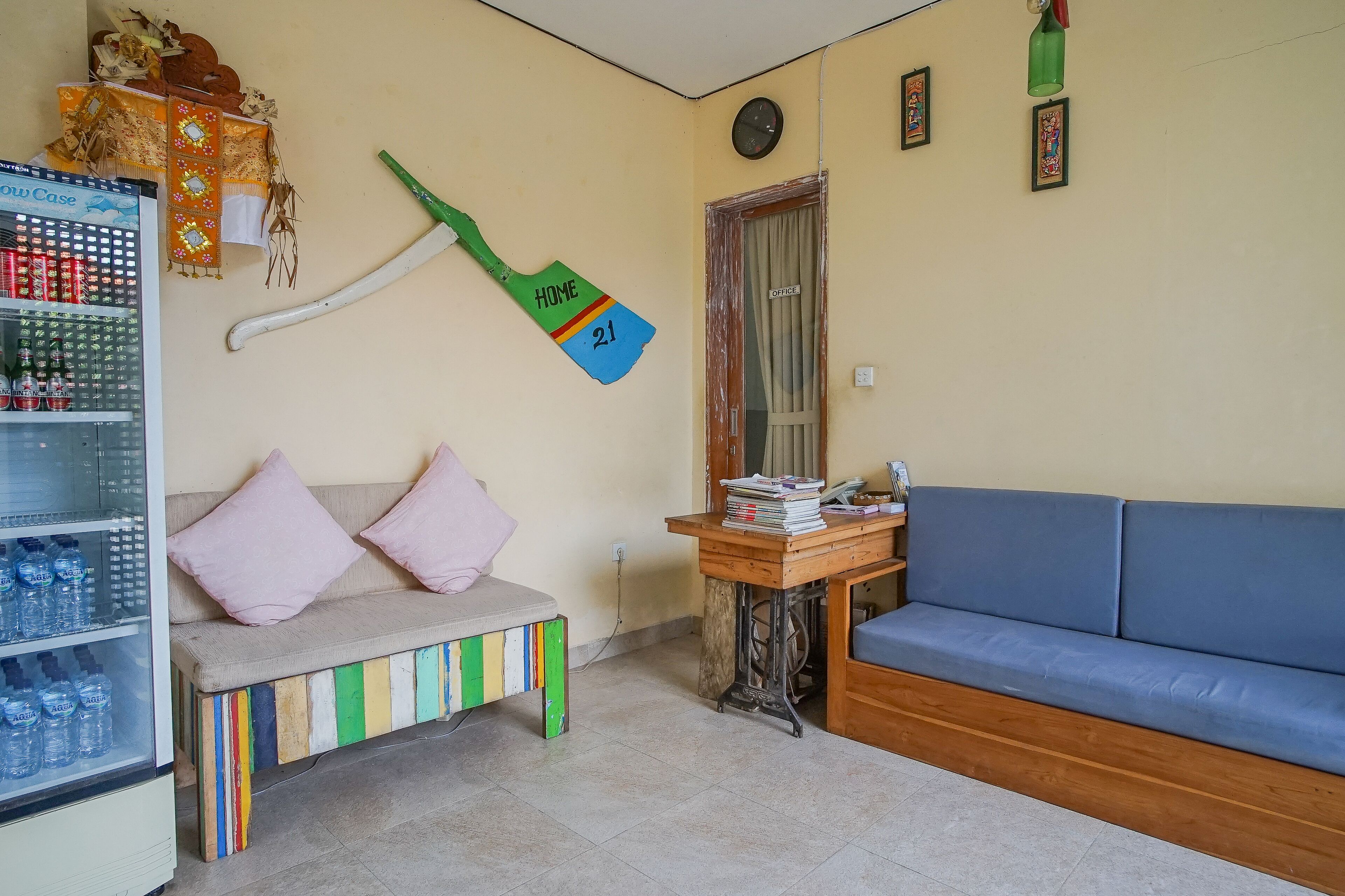 A room with a blue sofa, a wooden bench with colorful stripes, a refrigerator stocked with beverages, and a wall-mounted green and blue sign reading "HOME 21".
