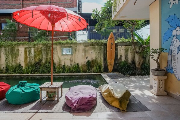 Uma área à beira da piscina com um guarda-sol vermelho, almofadas de feijão verdes e vermelhas, e uma prancha de surf encostada à parede.