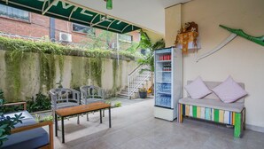 Un patio con mesa y sillas de madera, un banco colorido, un refrigerador lleno de bebidas y una escalera que lleva a un nivel superior.
