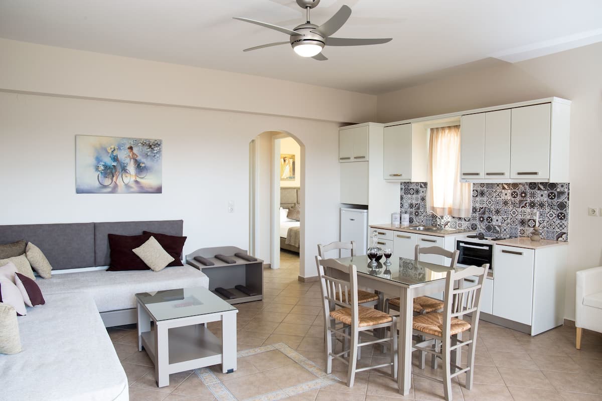A modern living room with a sofa, coffee table, and dining area. There is a kitchen with white cabinets and a tiled backsplash. A ceiling fan is visible, and a painting hangs on the wall.