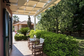 A covered patio with a table and chairs, surrounded by greenery and a stone wall. - Dina Apartments (Kefalonia)