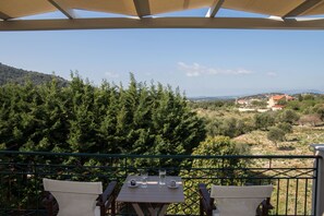 A terrace with a table and chairs, overlooking a mountainous landscape with trees and buildings. - Dina Apartments (Kefalonia)
