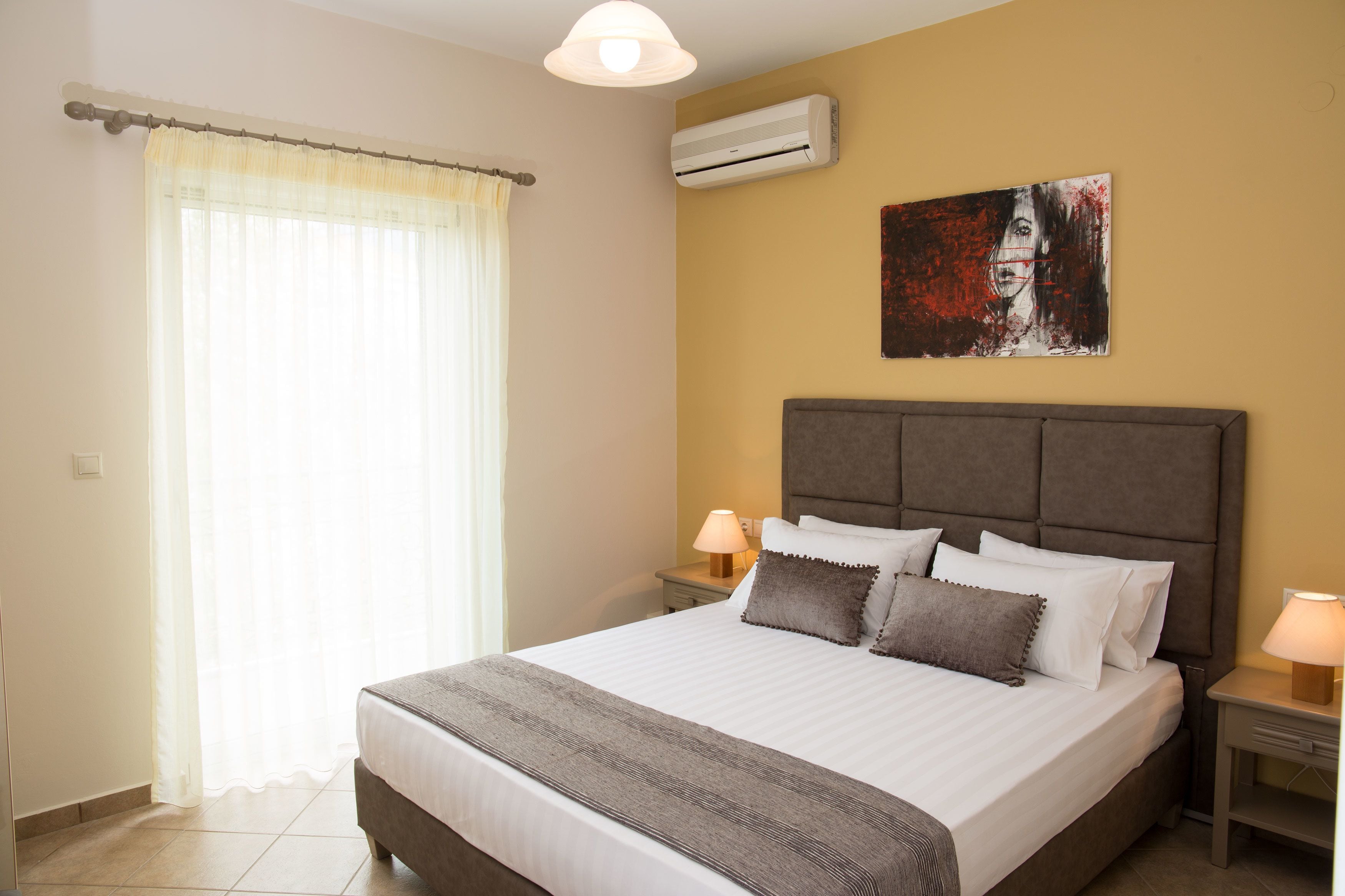 A hotel room with a large bed, two bedside tables with lamps, a wall-mounted air conditioner, and a painting above the bed.