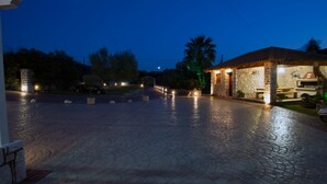 Un camino pavimentado que conduce a un edificio de piedra con un espacio al aire libre cubierto, posiblemente un patio o sala de estar, y una chimenea de piedra.