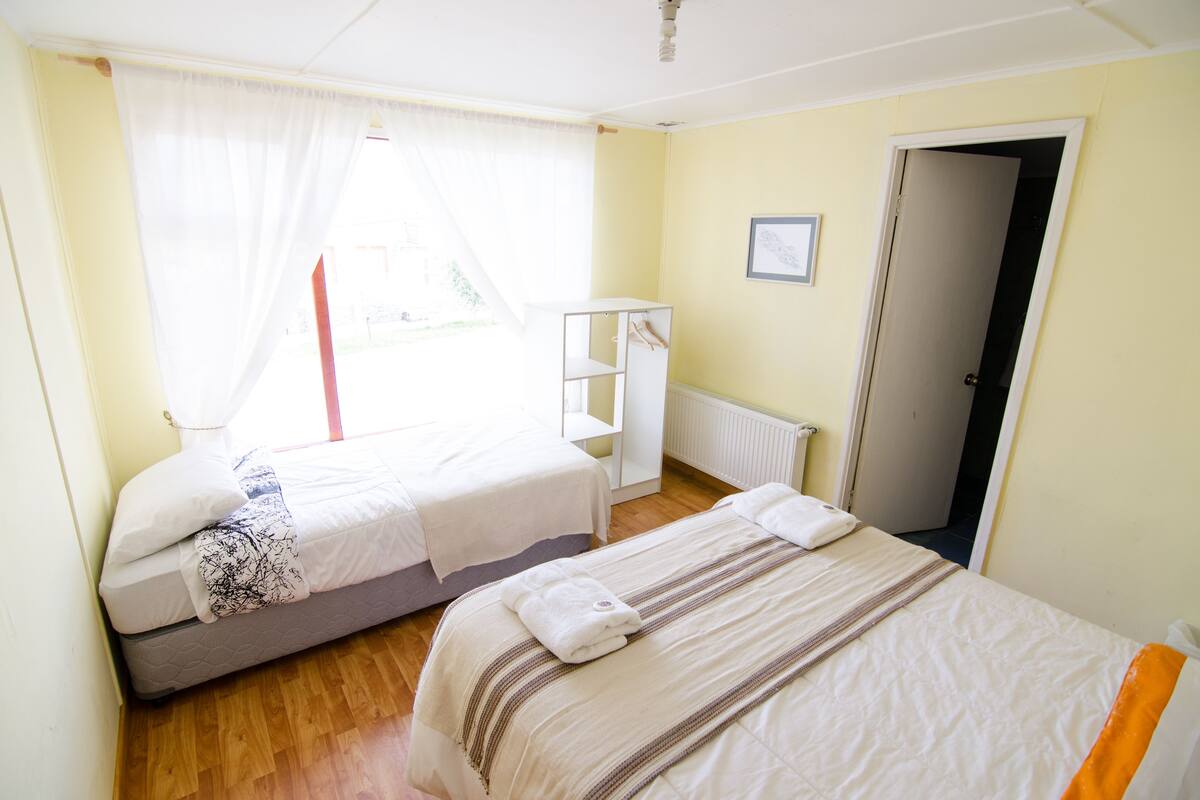 A bedroom with two beds, a window with sheer curtains, a radiator, and a door leading to another room.
