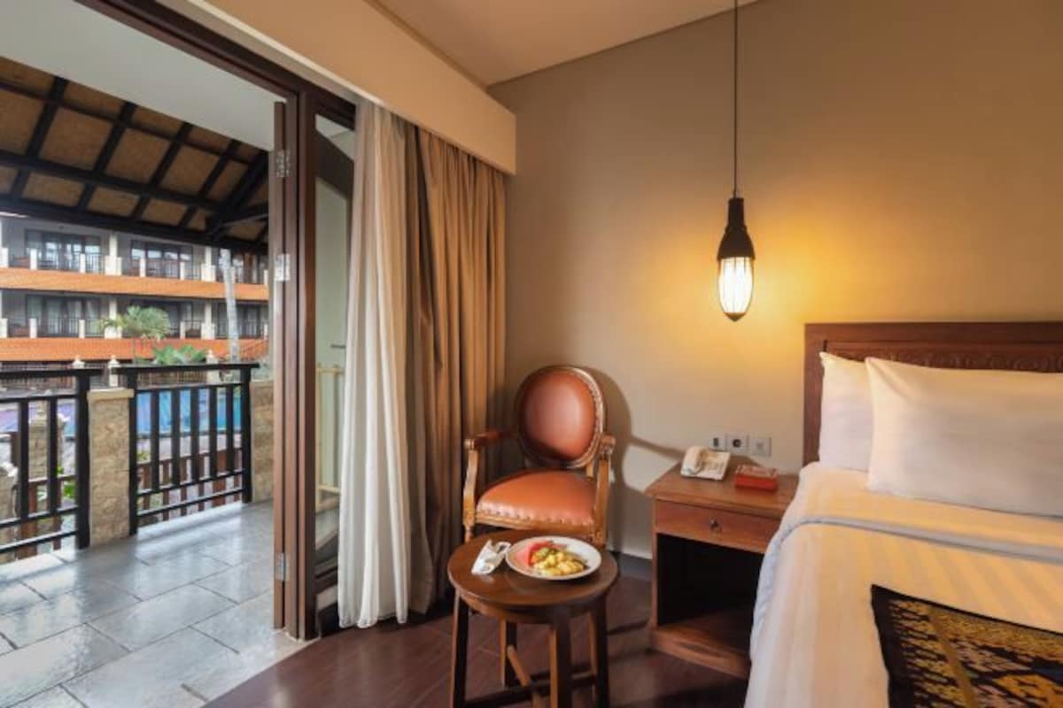 A hotel room with a bed, a chair, a table with a plate of fruit, and a balcony with a view of other buildings.