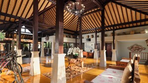 A spacious lobby with a high ceiling, wooden beams, and a chandelier. There are bicycles parked against the wall, a wooden bench with cushions, and a table with a vase of flowers. - Best Western Premier Agung Resort Ubud (Ubud)