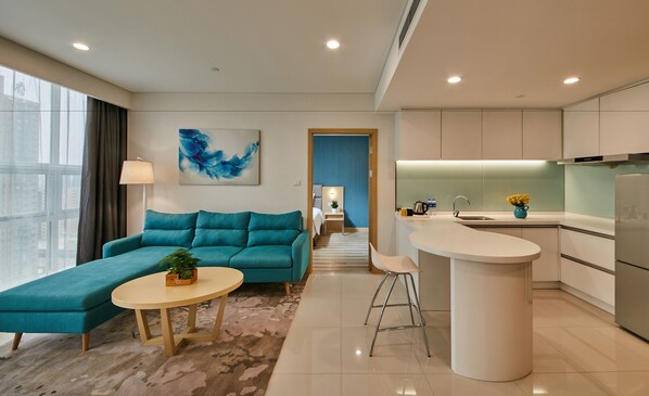 A modern living room with a teal sofa, a small round table, and a kitchen area with a white countertop and stainless steel appliances. - Holiday Inn Express Luoyang Yichuan by IHG (Luoyang)
