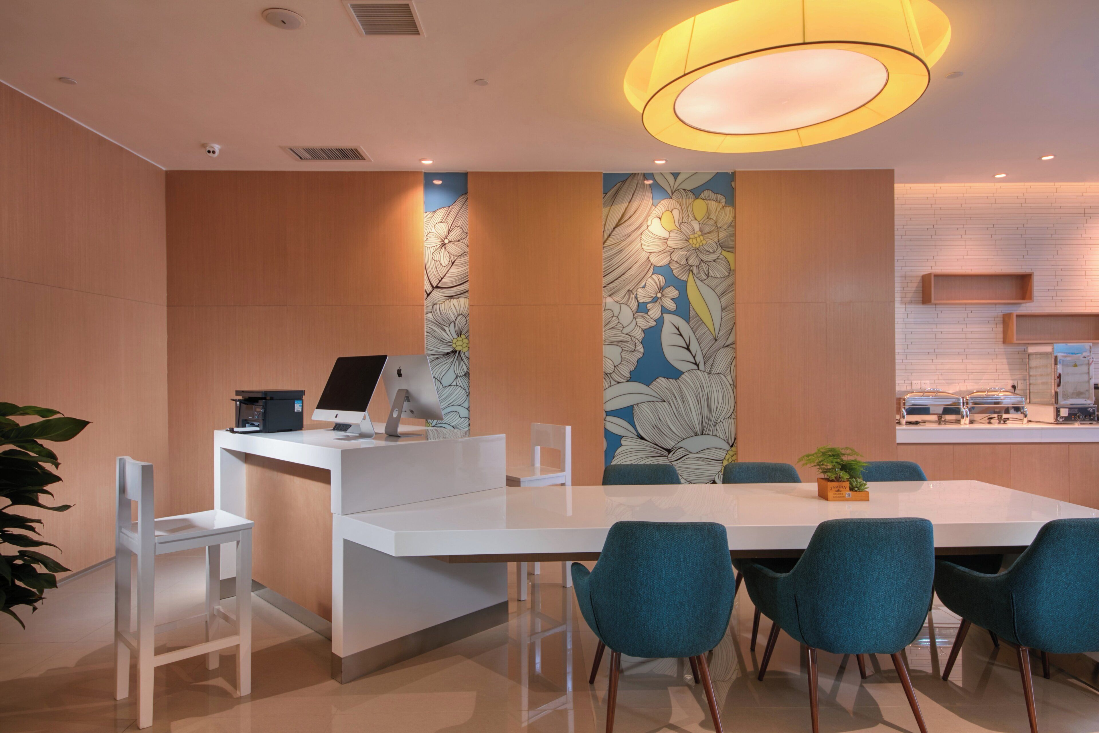 A modern meeting room with a white desk, two Apple iMac computers, teal chairs, a large floral wall mural, and a kitchen area in the background.