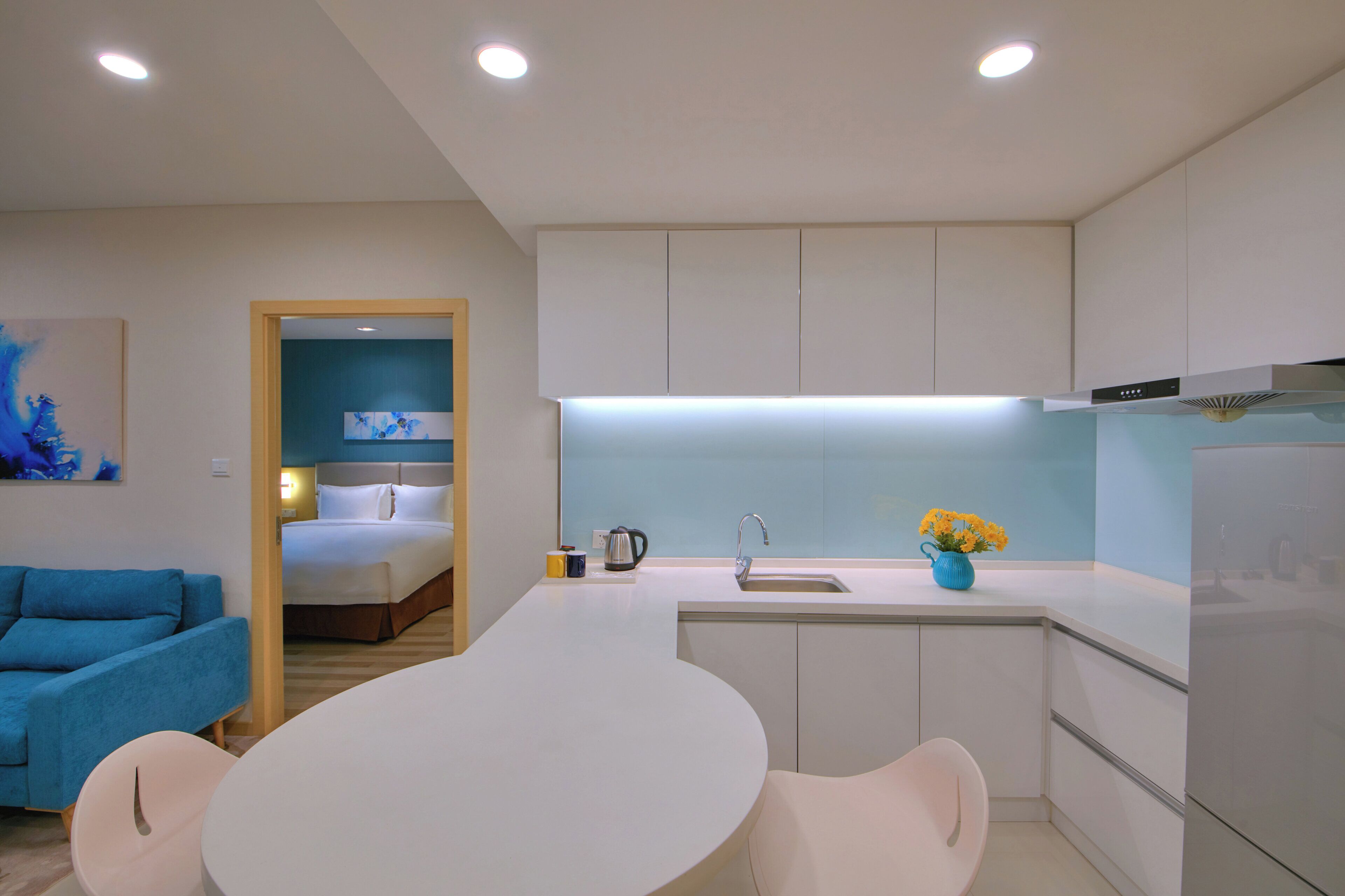 A modern kitchen with a central island, a dining area with a table and chairs, and a bedroom visible through an open door.