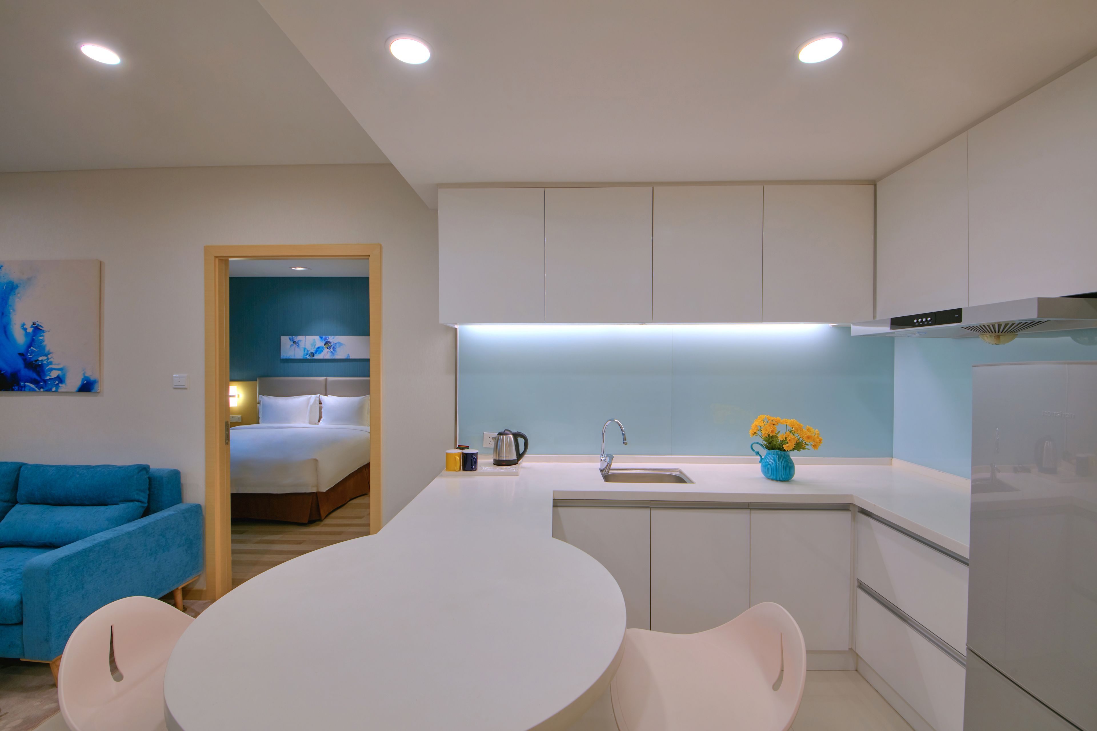 Una cocina moderna con armarios blancos, una isla central con asientos, un horno empotrado y vista a un dormitorio.