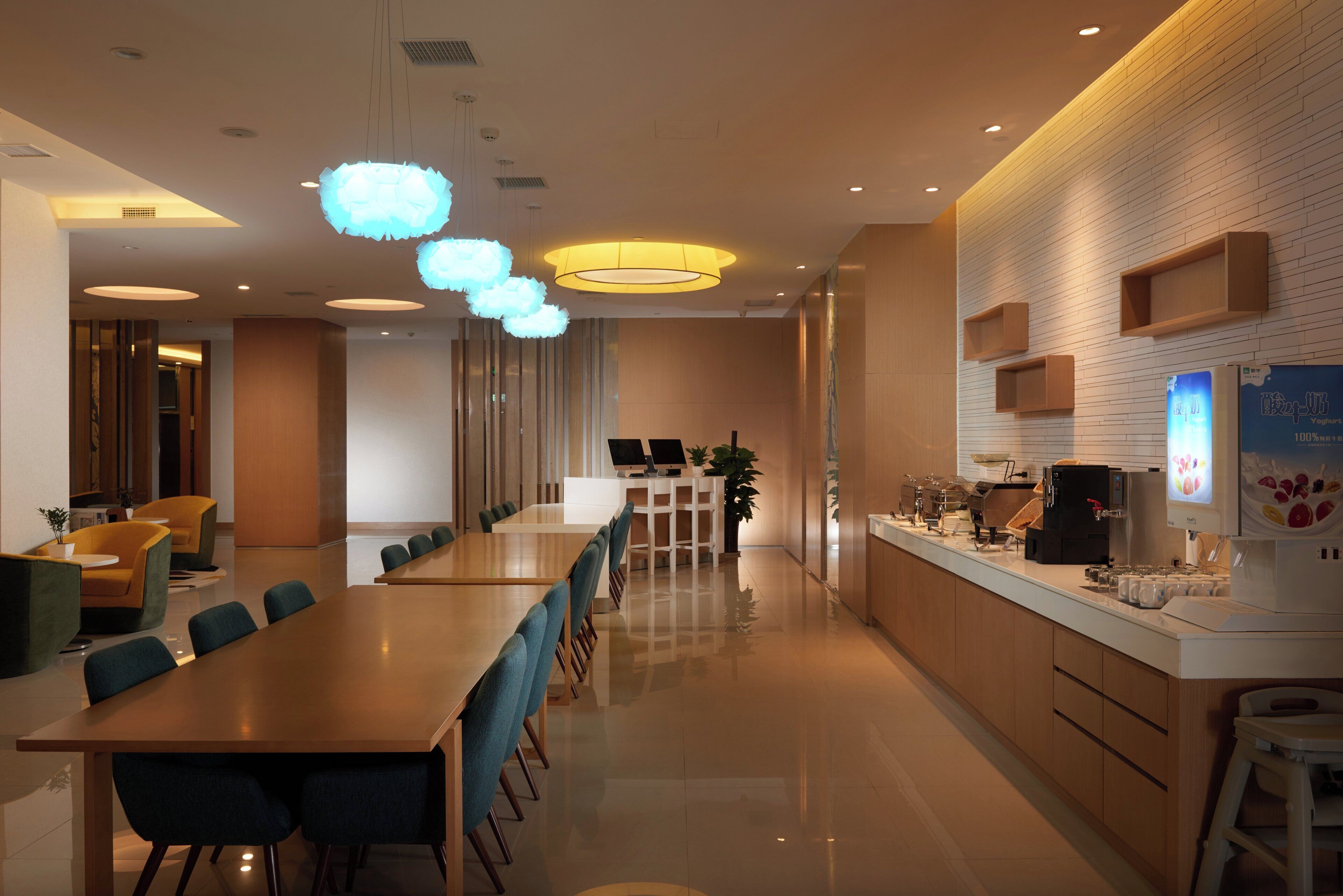 A modern hotel lobby with a long reception desk, seating area, and a kitchen counter with coffee machines.