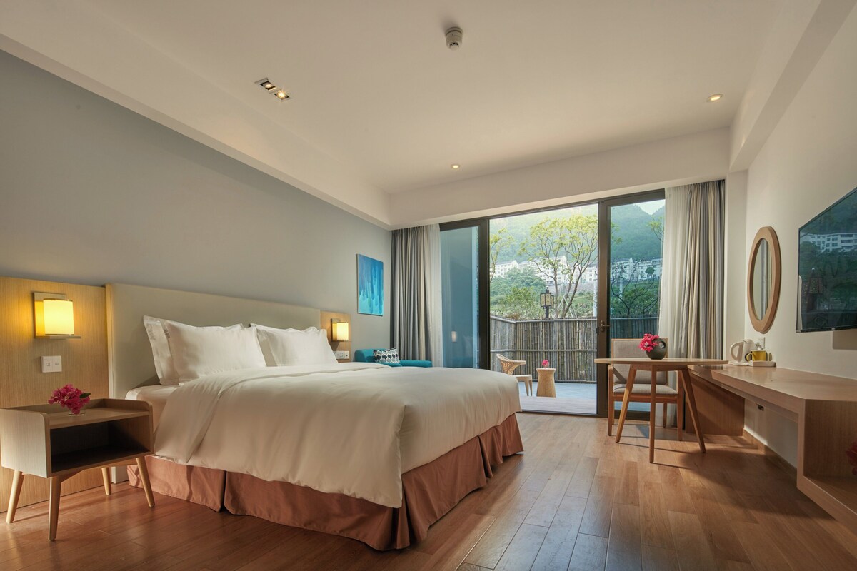 A hotel room with a large bed, a desk, a chair, a TV, and a balcony with a view of the city.