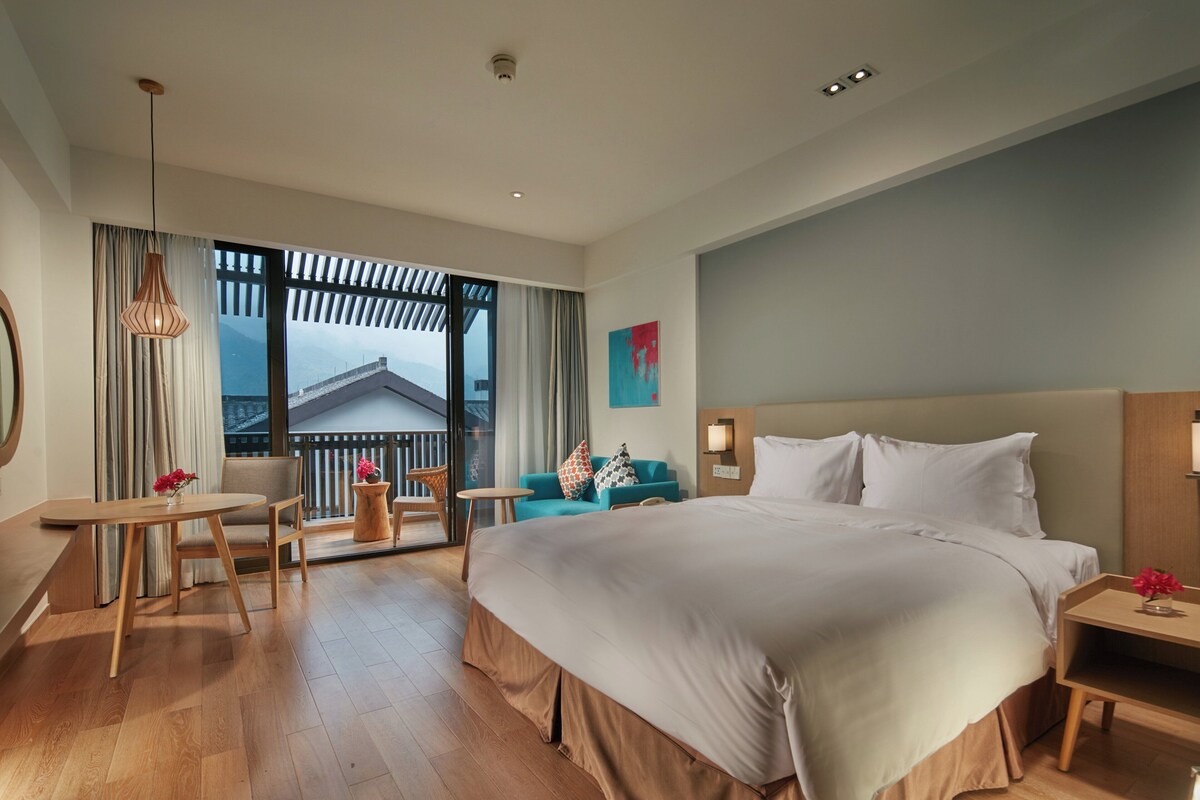 A hotel room with a large bed, a sitting area with a table and chairs, a view through a large window, and a ceiling with recessed lighting.