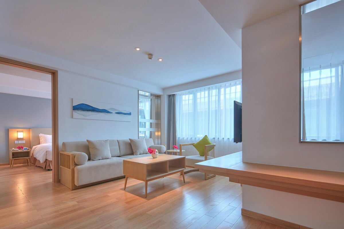 A modern hotel room with a sofa, a coffee table, a TV, and a large window with curtains.