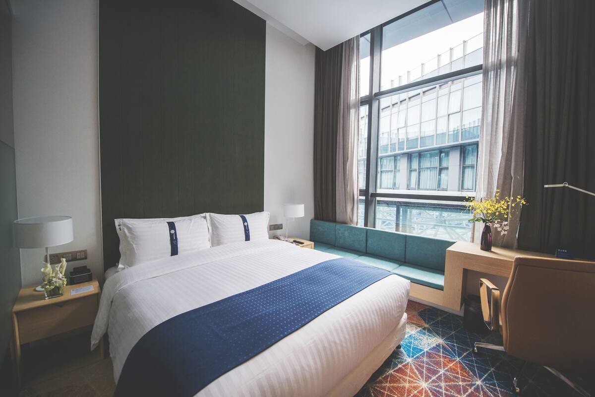 A hotel room with a large bed, a desk, a chair, a nightstand, a lamp, and a window with curtains.