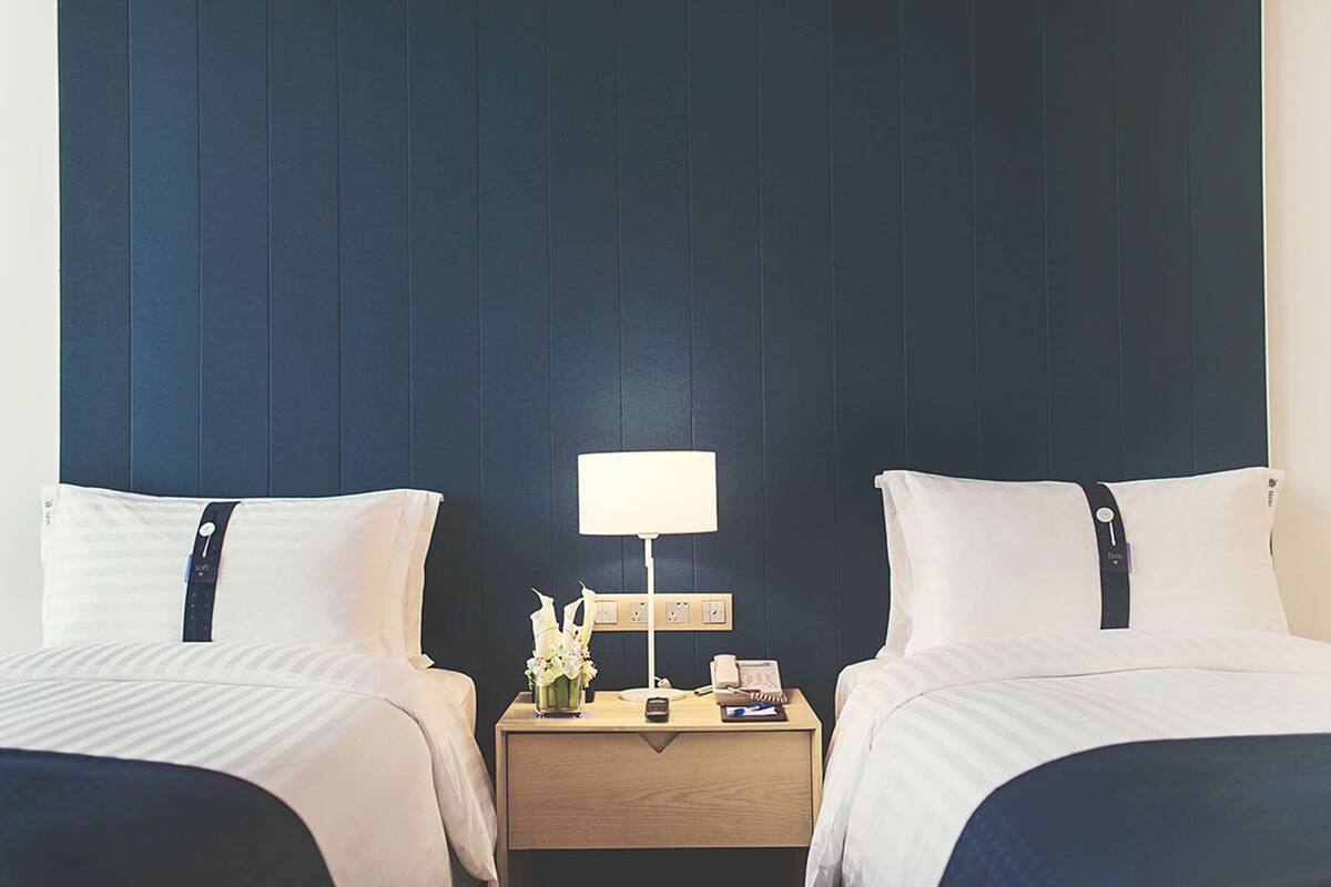 A hotel room with two beds, a nightstand with a lamp, a phone, and a small vase with flowers.