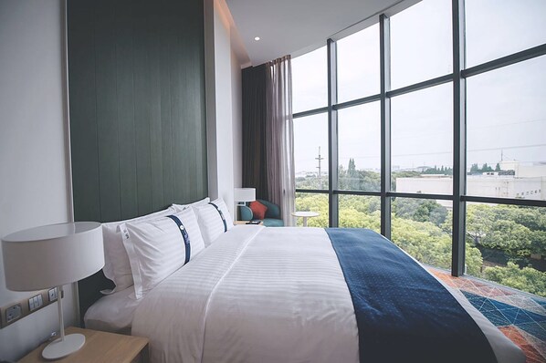 A hotel room with a large bed, a view of greenery and buildings through the window, and a modern design.