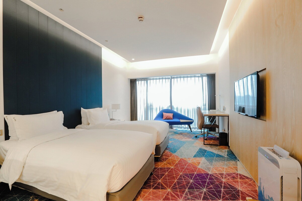 A hotel room with a large bed, a desk with a chair, a TV, and a window with curtains.
