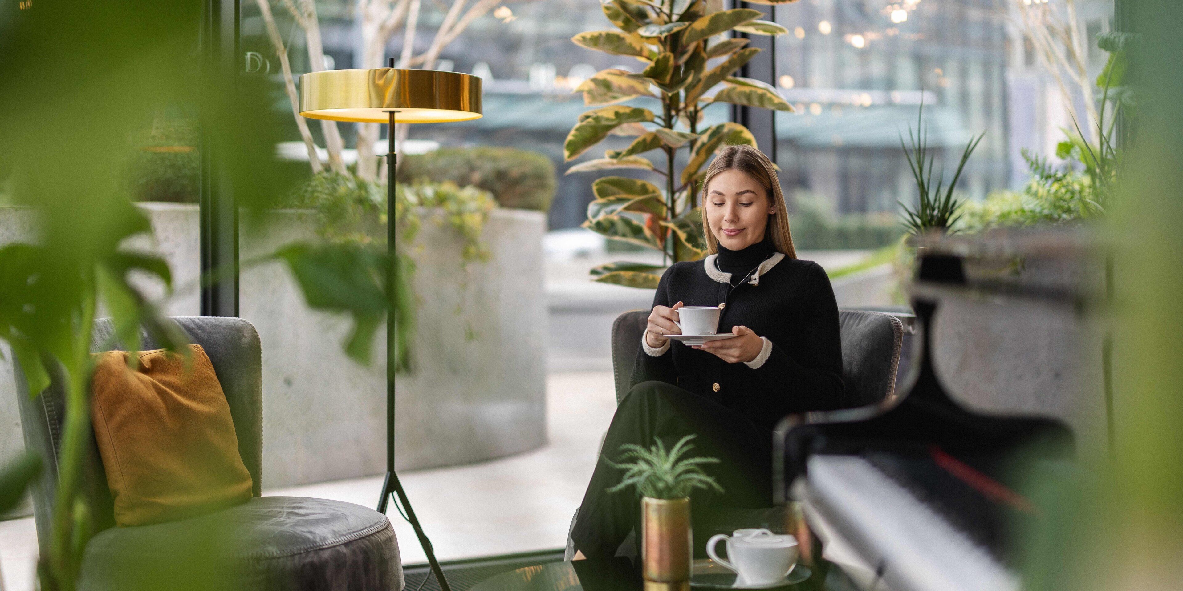 A person seated on a couch in a modern indoor space with a floor lamp, a coffee cup, and a potted plant.