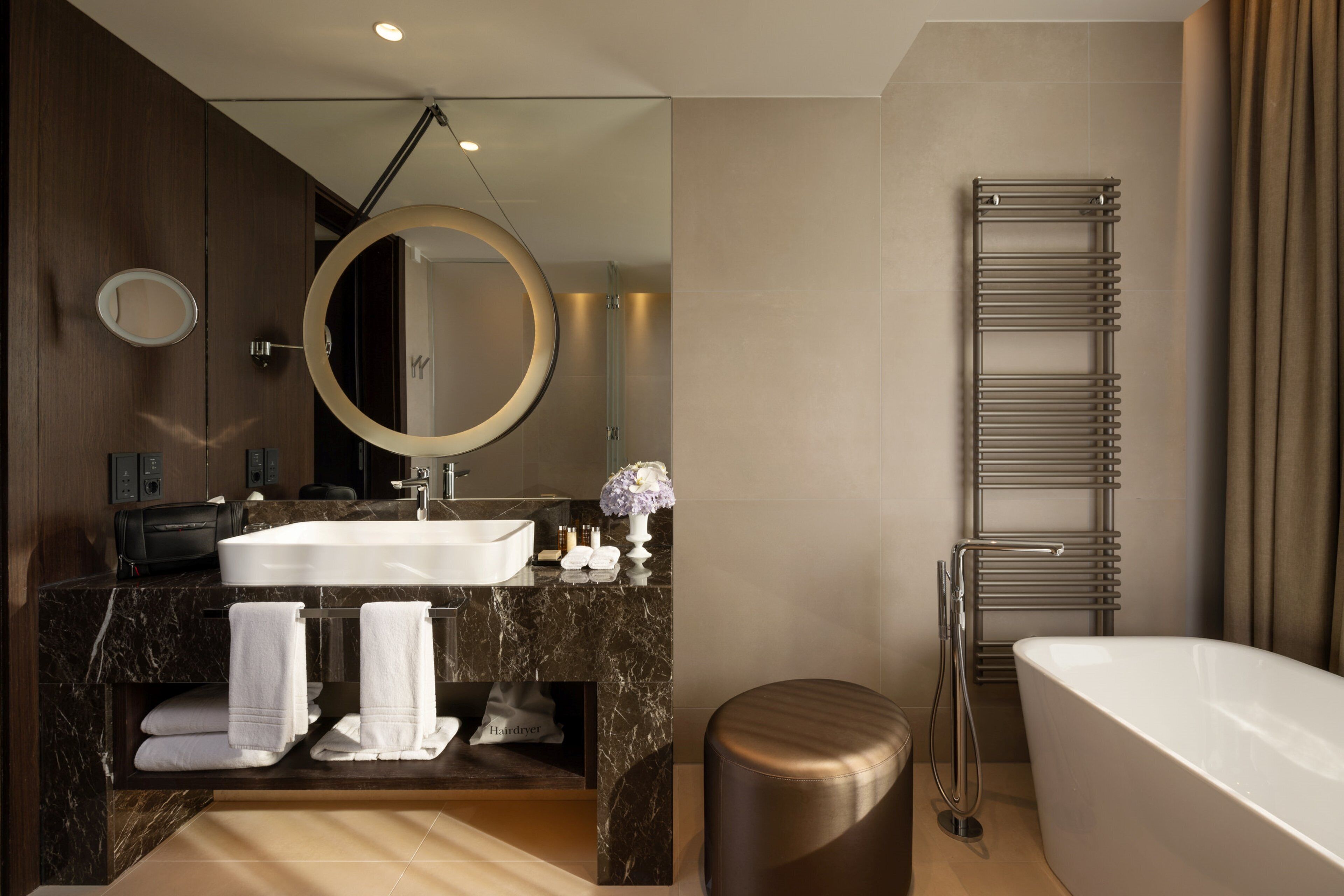 A modern bathroom with a large bathtub, a round mirror, a built-in sink, and a towel rack.