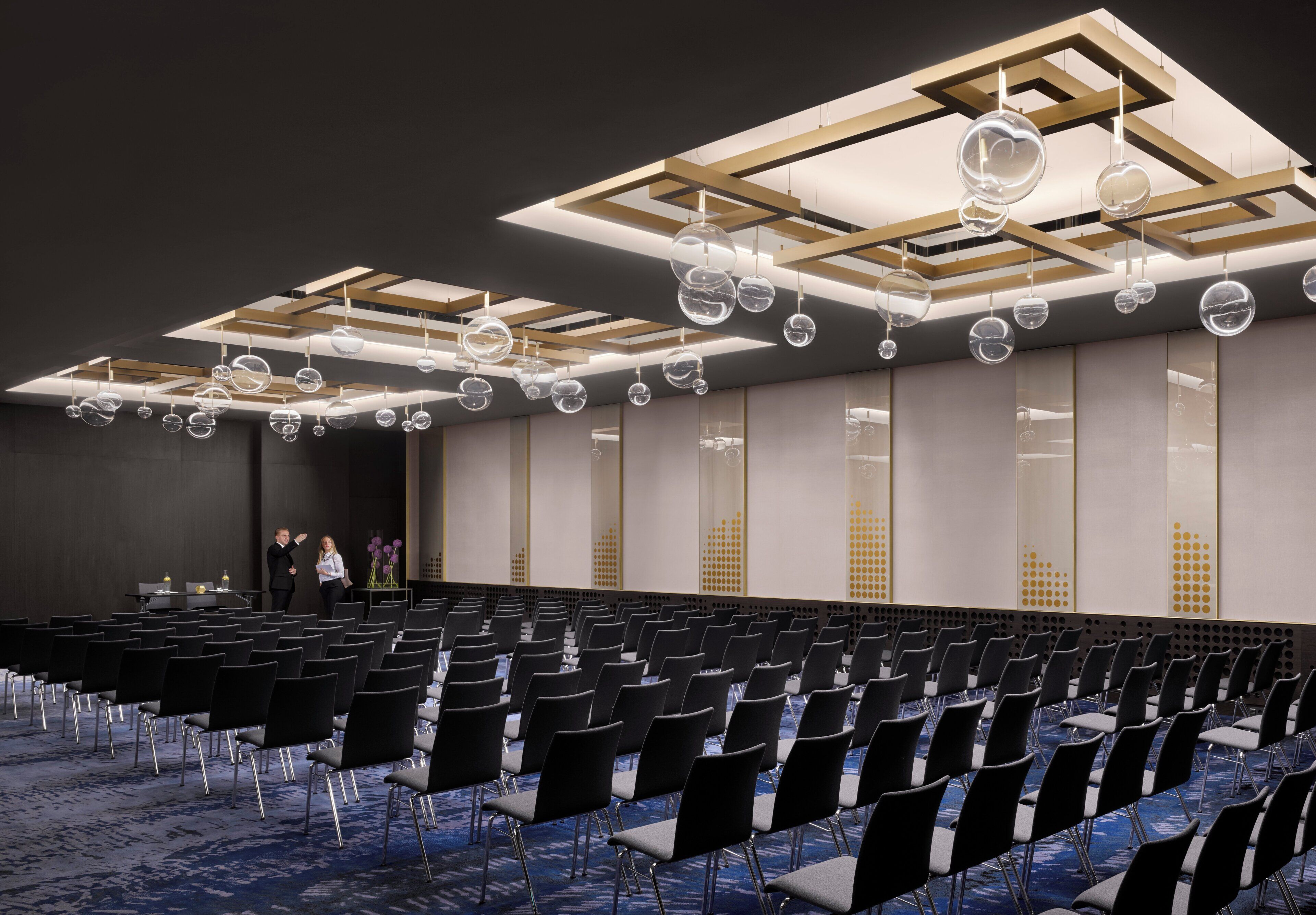A conference room with rows of chairs, a stage, and a ceiling with hanging spherical light fixtures.