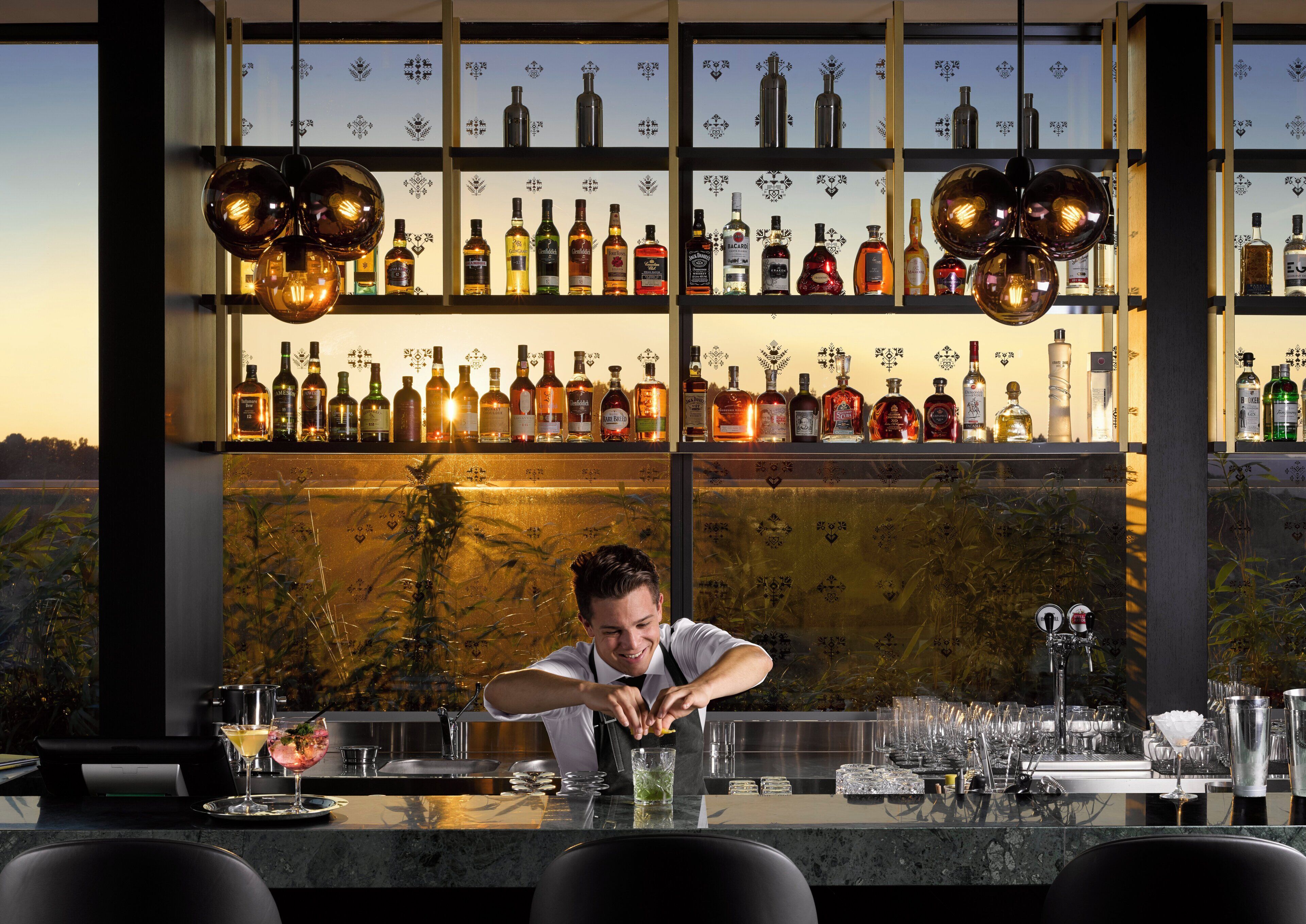Un espace bar avec un barman préparant des boissons, des étagères remplies de bouteilles d’alcool et une vue sur l’extérieur.