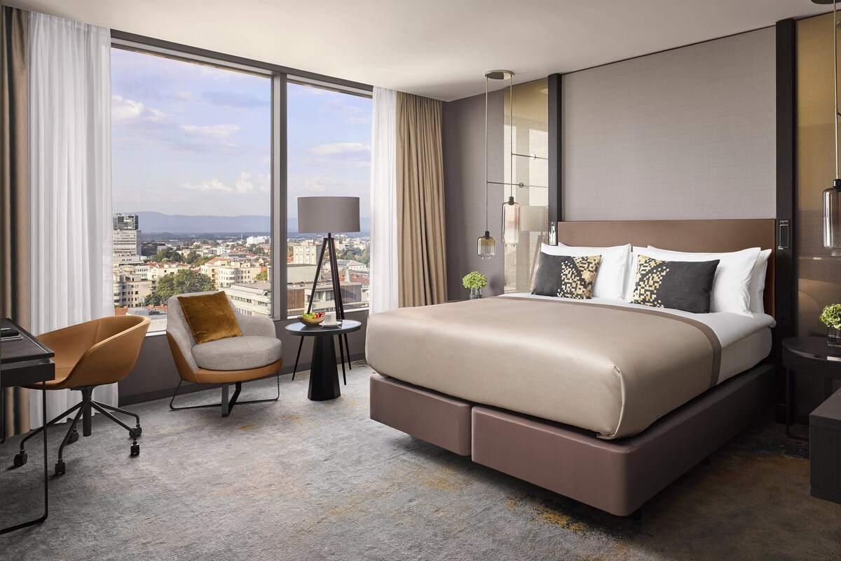 Une chambre d’hôtel moderne, dotée d’un grand lit, d’un bureau, d’une chaise et offrant une vue sur la ville.