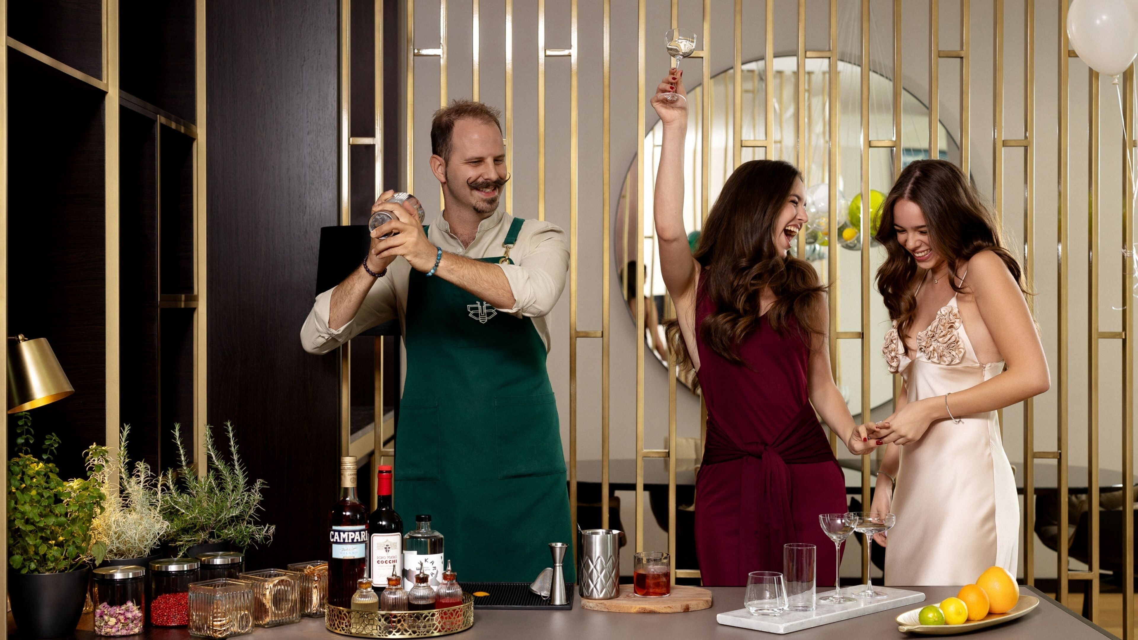 A bar area with a bartender preparing drinks, a woman holding a glass, and a man with a drink. There are bottles, glasses, and a fruit plate on the counter.
