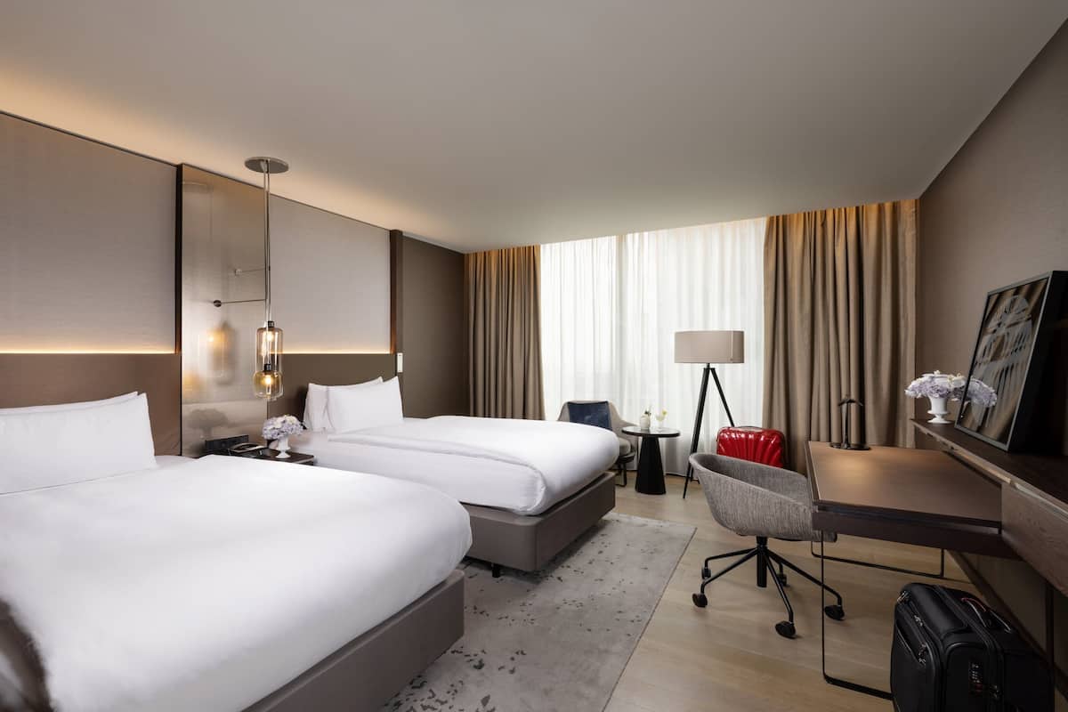 Een hotelkamer met twee bedden, een bureau, een stoel, een lamp, een koffer en een schilderij aan de muur.