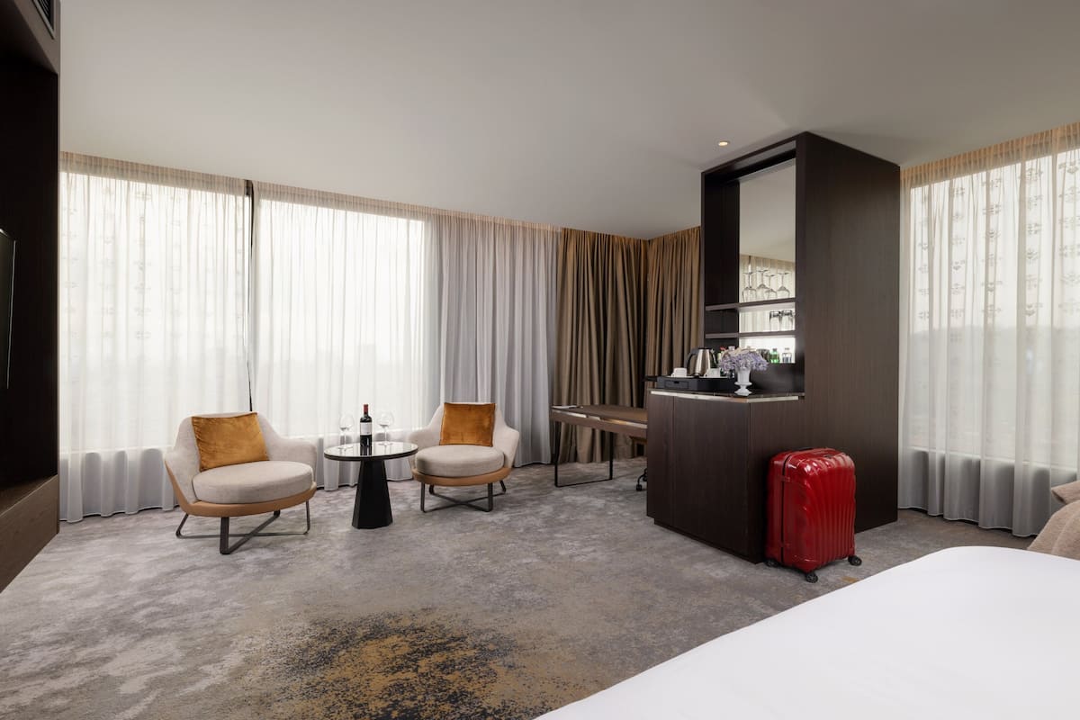 Een hotelkamer met een bed, twee fauteuils, een kleine tafel met een fles wijn, een rode koffer en een minibar.
