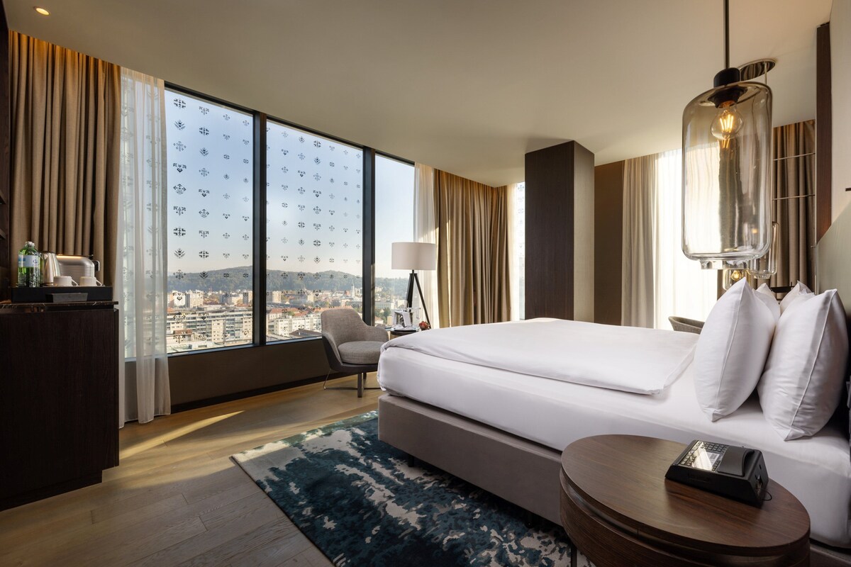 Une chambre d’hôtel moderne dotée d’un grand lit, d’un bureau, d’une chaise, d’une lampe et offrant une vue sur la ville.
