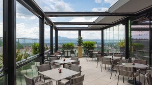 Una terraza en la azotea con paredes de cristal, mobiliario exterior y vistas a la ciudad y montañas.