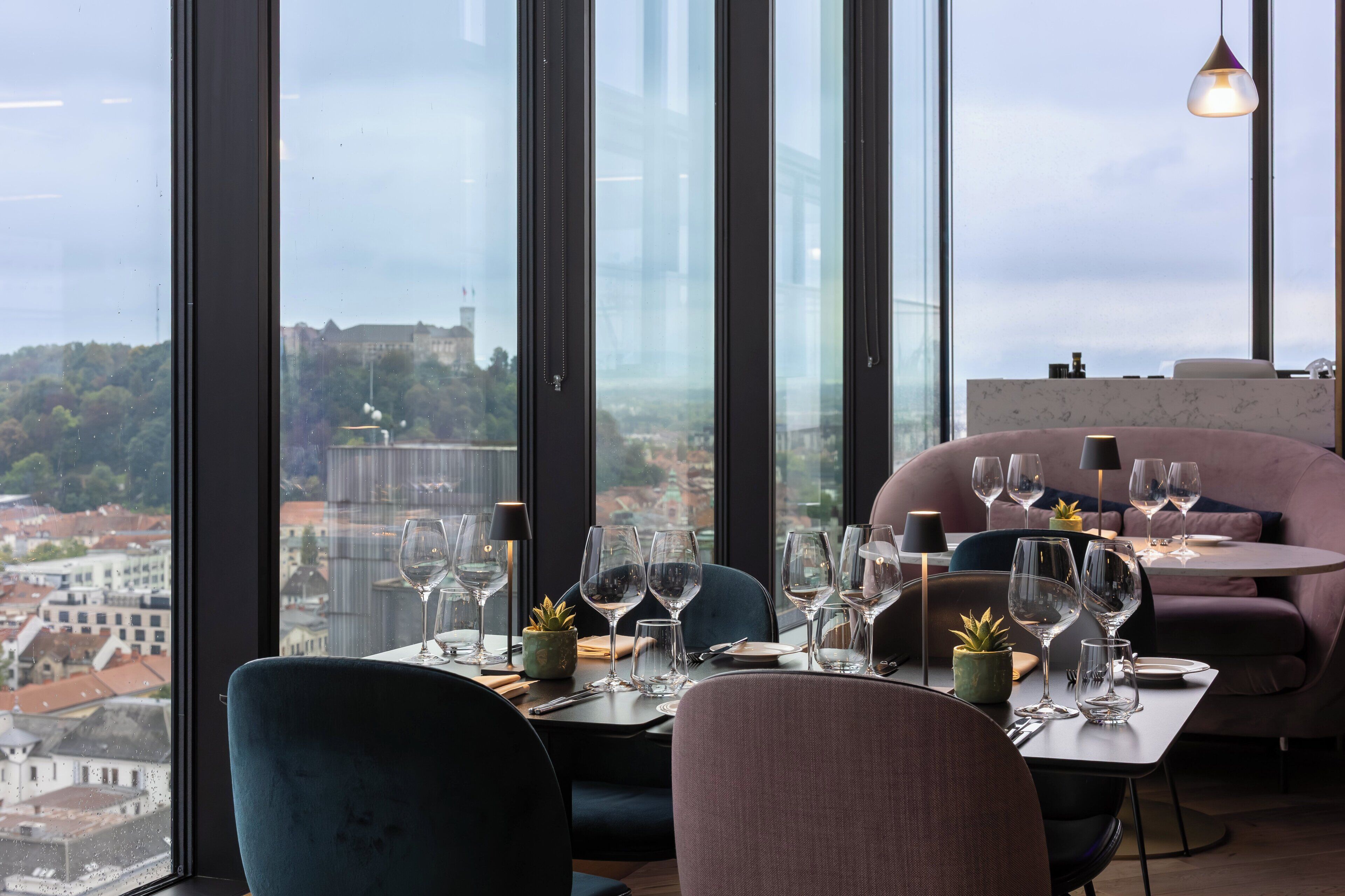 A dining area with a table set for a meal, large windows offering a city view, and a cozy seating area with cushions.