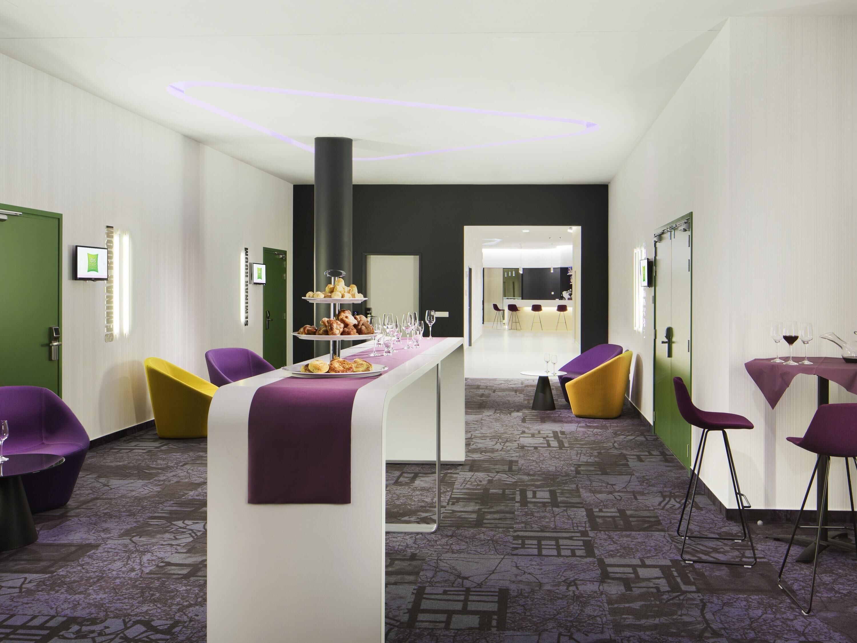 A modern hotel lobby with a reception desk, purple and yellow chairs, a buffet table, and a bar area.