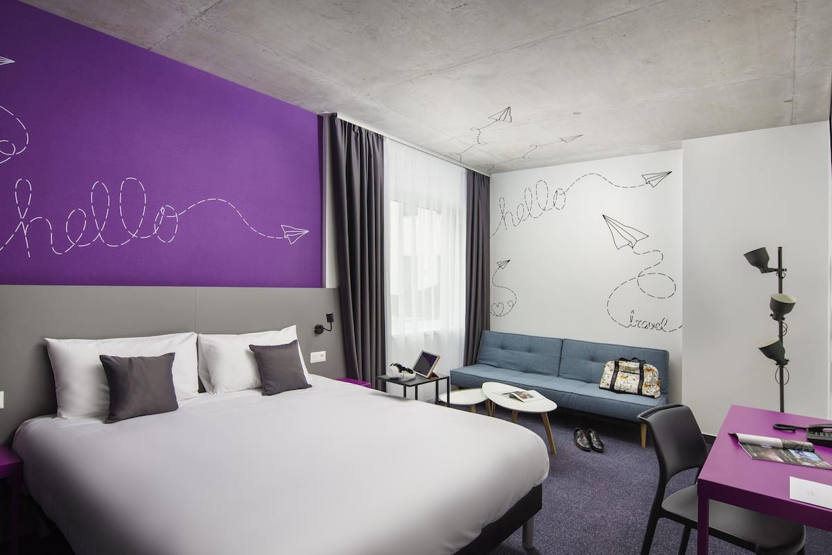 Een moderne hotelkamer met een groot bed, een zithoek, een bureau en een paarse accentmuur met tekeningen over reizen.
