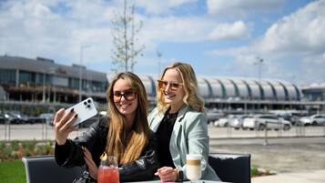 Dos personas sentadas en una mesa al aire libre, con una bebida y un cuaderno, y un edificio moderno de fondo.