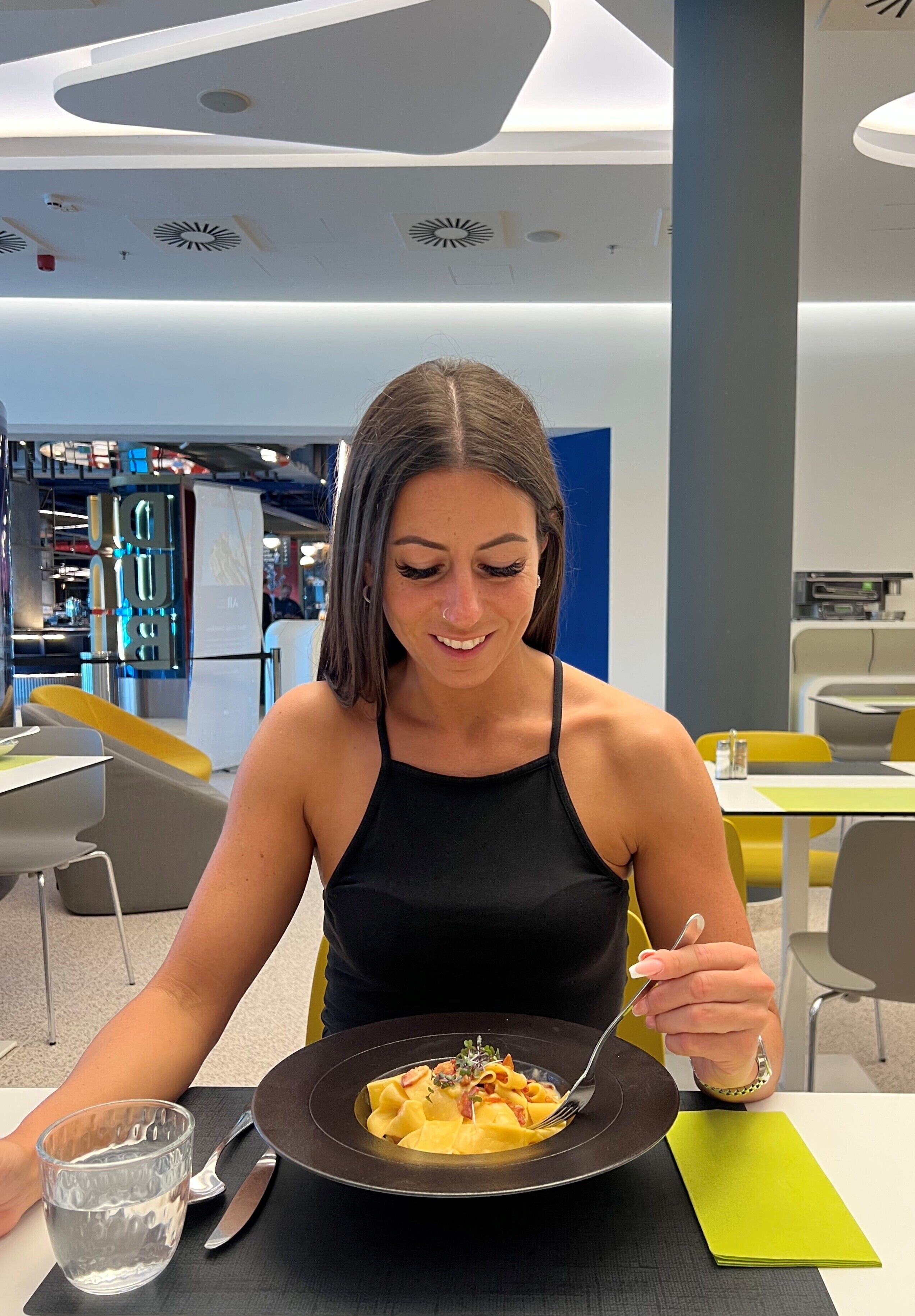 Una persona che cena in un ristorante dall'arredamento moderno, con un piatto nero pieno di cibo, un bicchiere d'acqua e una forchetta sul tavolo.
